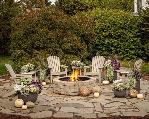 Outdoor fire pit surrounded by Adirondack chairs and decorative plants.