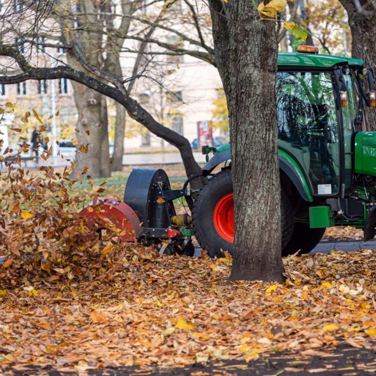 Green tractor clearing fallen leaves in a park during autumn.